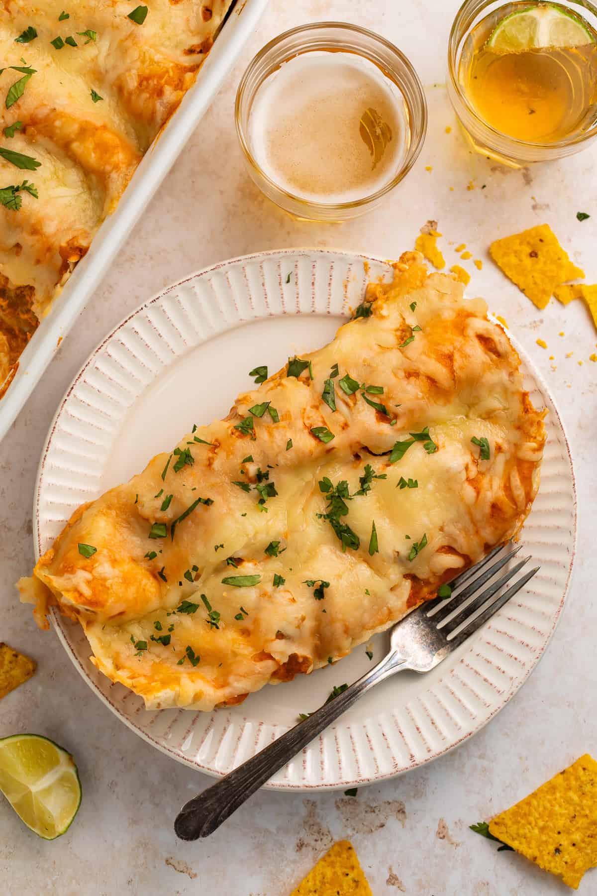 Two chicken enchiladas on a white plate with a form,. There are glasses of beer and the pan of other enchiladas off to one side.