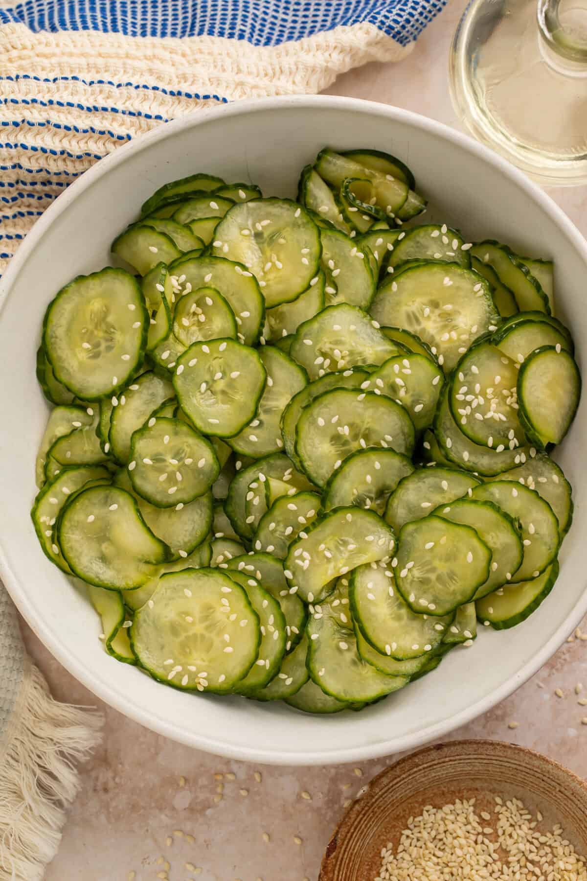 White bowl of Asian cucumber salad with toasted sesame seeds. A blue and white kitchen towel, a bottle of vinegar and a wooden bowl of sesame seeds is pictured off to one side.