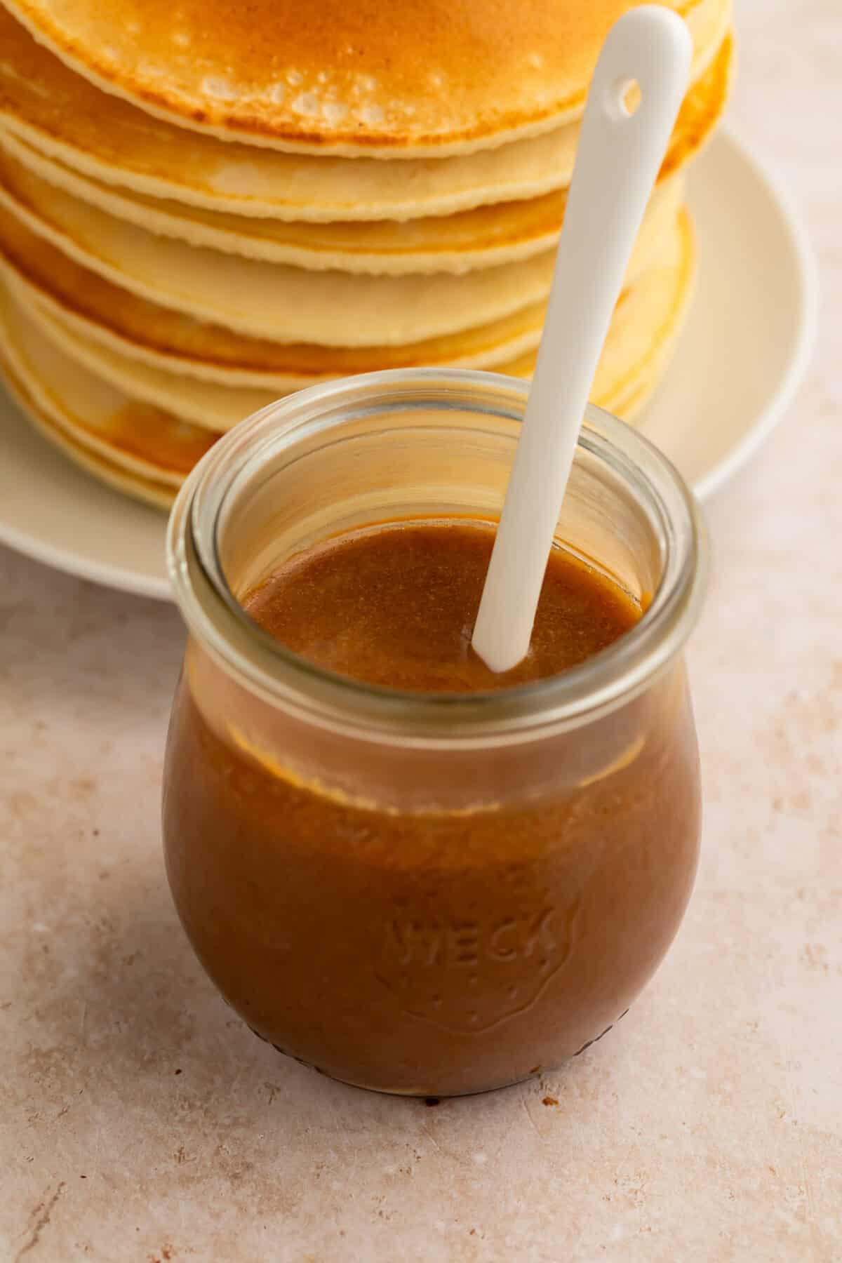A jar of peanut butter pancake syrup with a white spoon in front of a stack of buttermilk pancakes.
