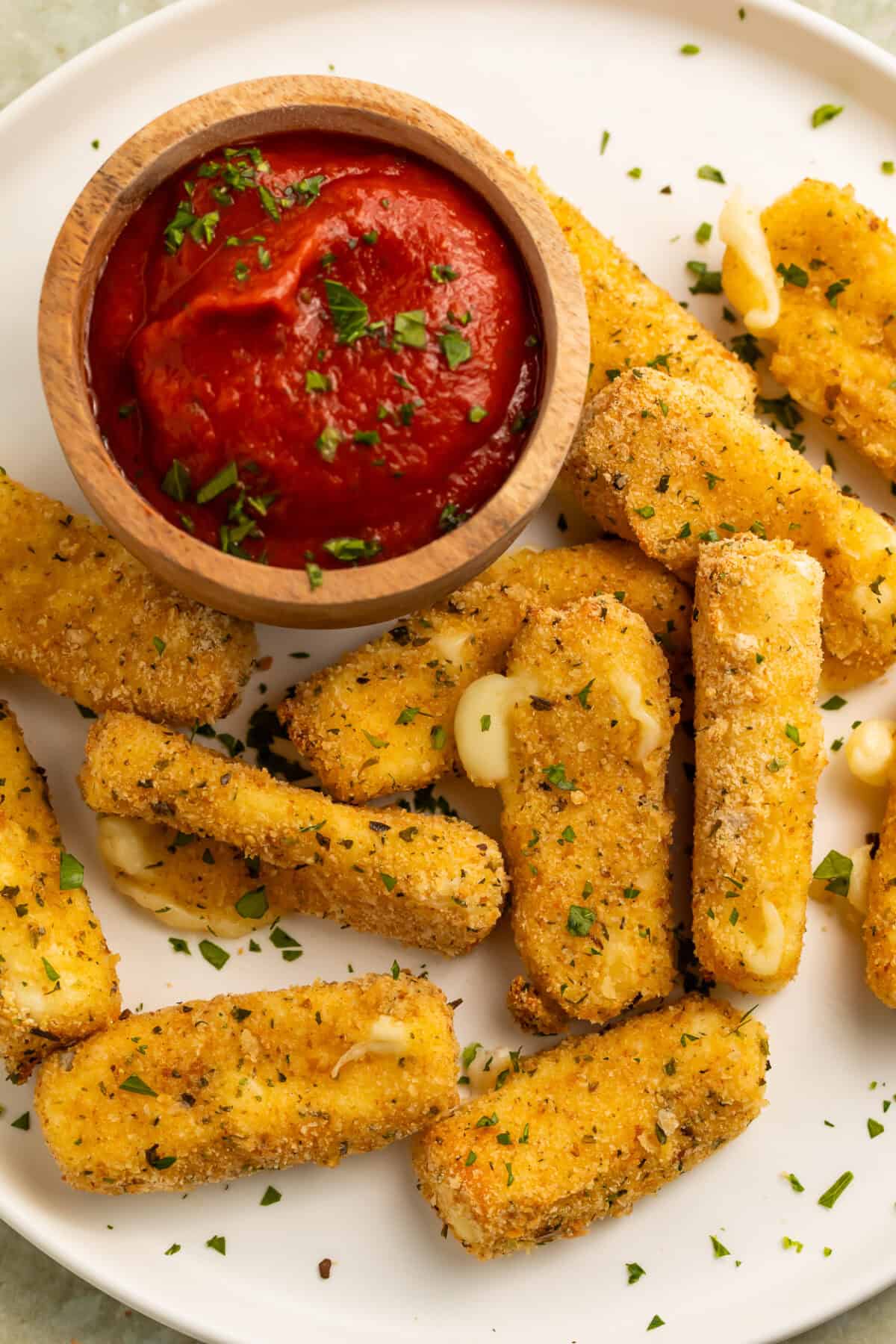 Close up of a white plate of air fryer mozzarella sticks with a scattering of finely chopped herbs and a wooden bowl of tomato dipping sauce.
