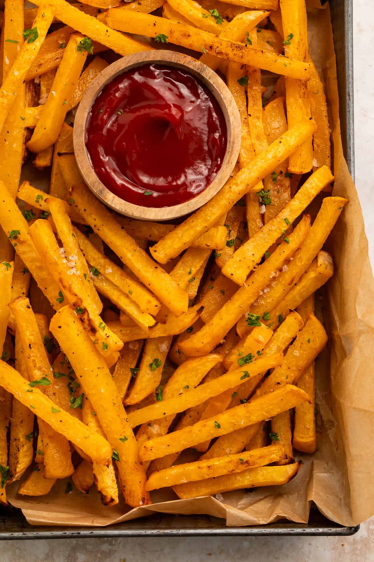 Close up of a baking parchment-lined tray with rutabaga fries and a wooden pot of ketchup.