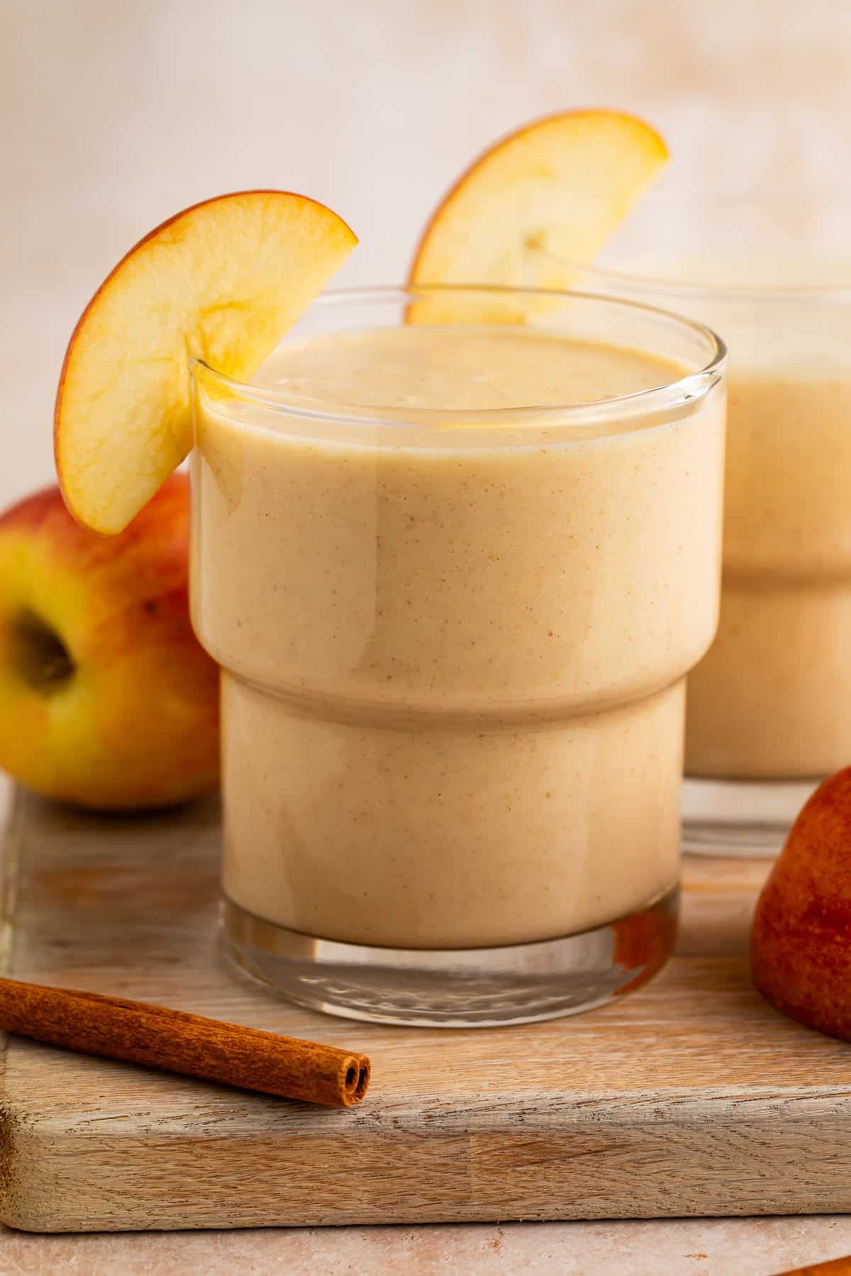 Side on view of an apple smoothie in a glass resting on a wooden board. There are apples and cinnamon sticks next to it.
