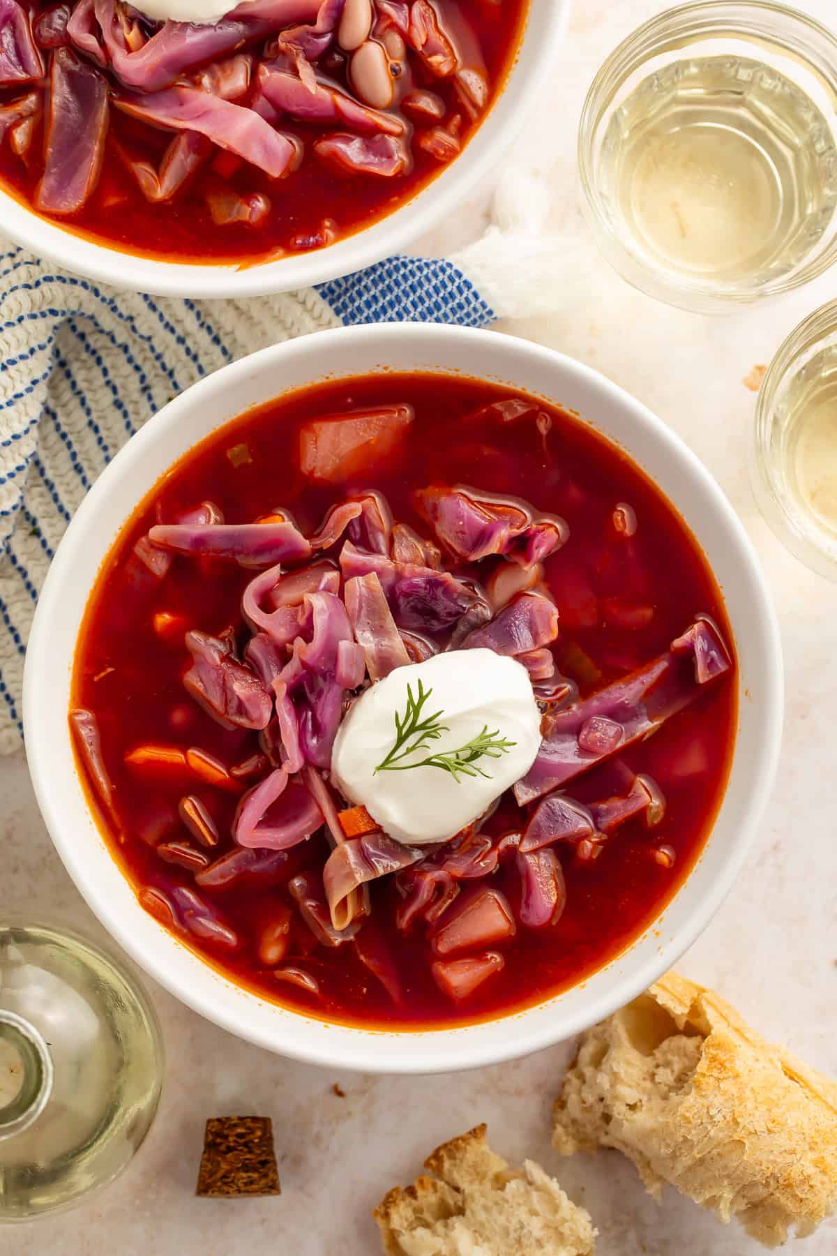 Bowls of red cabbage soup with a dollop of sour cream on top. Cups of wine and torn bread are pictured off to the side.