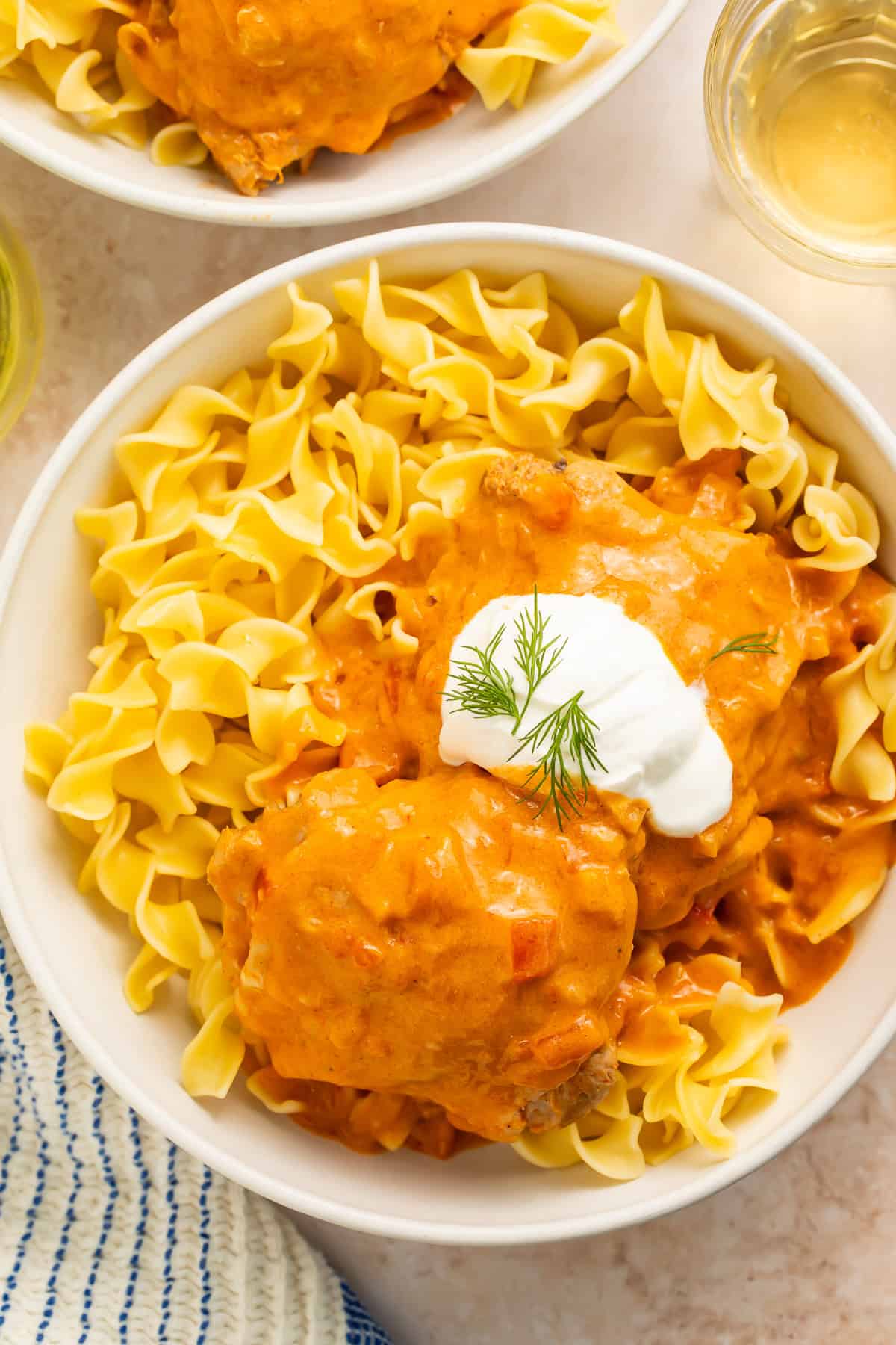 Bowl of creamy tomato and paprika chicken thighs served over egg noodles in a white bowl with a dollop of sour cream on top.