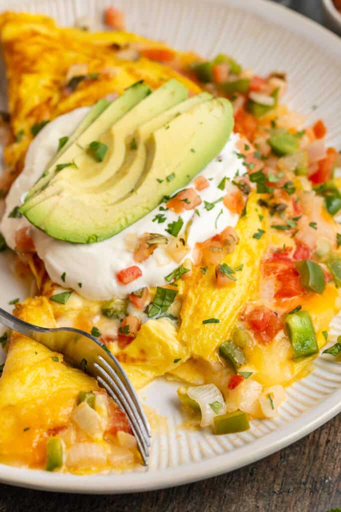 Angled view of a Mexican omelette filled with cheese, onion, and bell pepper, and topped with sour cream and slices of avocado.
