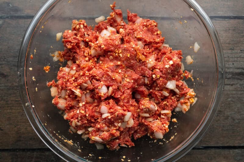 Overhead view of lean ground beef, diced onion, salt, pepper, minced garlic, and 1 large egg mixted together in a large glass mixing bowl.