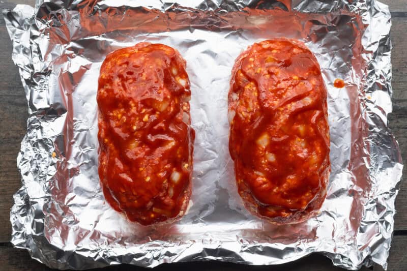 Overhead view of two uncooked meatloves on sheets of aluminum foil, topped with a ketchup glaze.