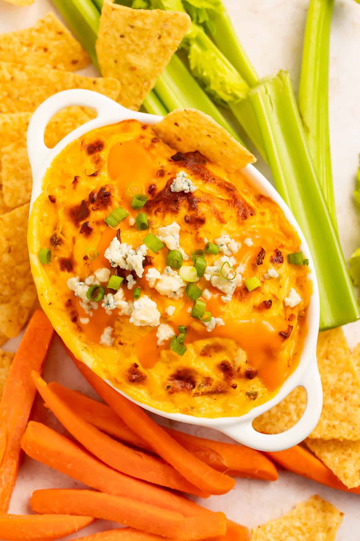 Overhead view of a high protein buffalo chicken tip, garnished with tortilla chips, crumbled cheese and sliced green onion. More tortilla chips, and carrot and celery sticks are arranged around the dish.