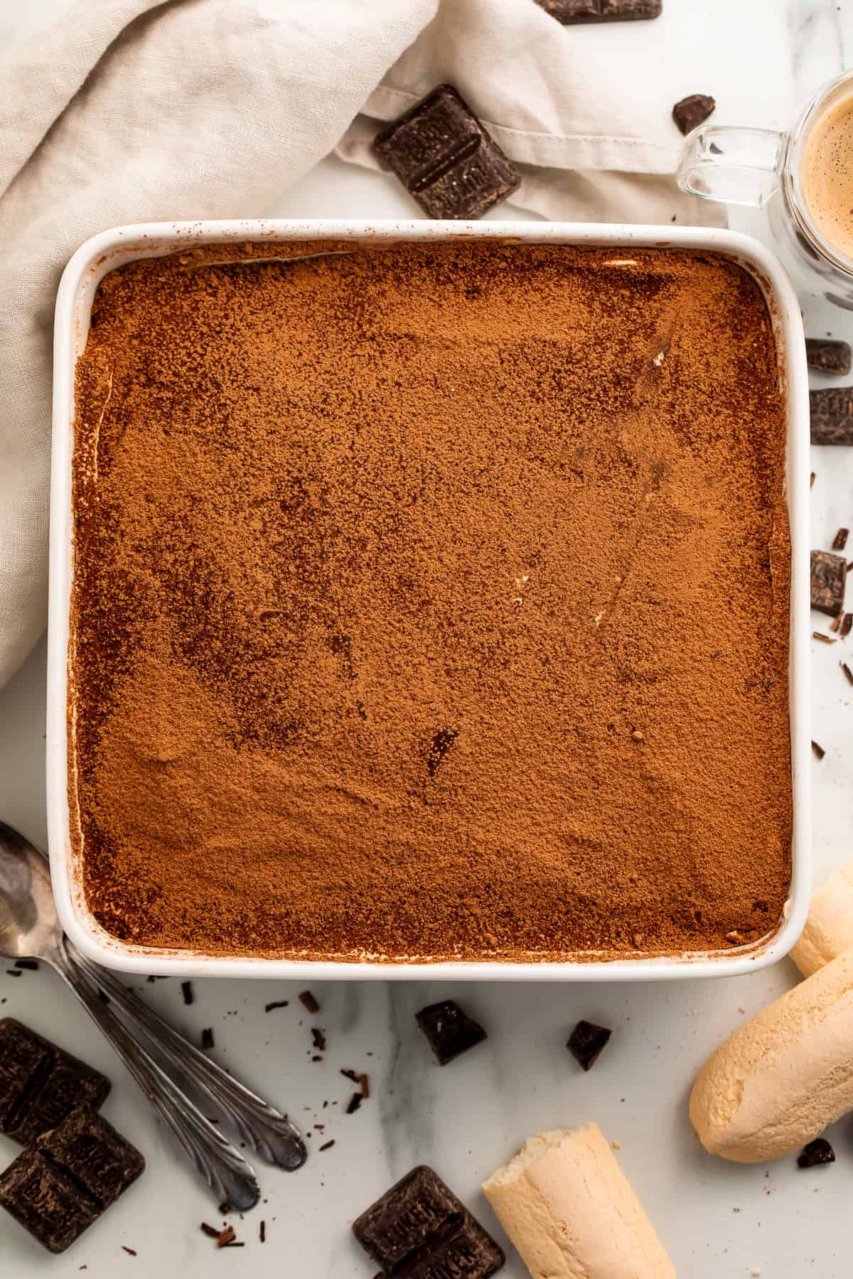 Overhead view of a gluten free tiramisu in a white baking dish with biscuits and dark chocolate squares scattered around. 