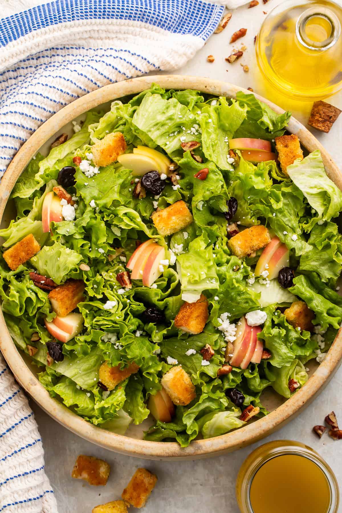 Wooden bowl of salad topped with apples and croutons, with a jug of yellow dressing on the side.