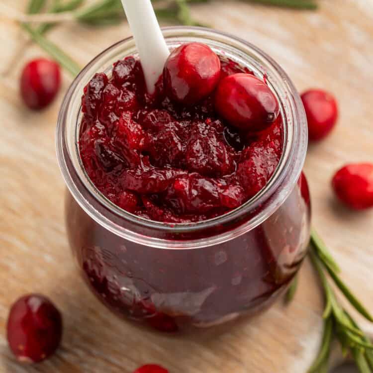 Jar of open cranberry jam with a white spoon on a wooden board scattered with rosemary sprigs and fresh cranberries.