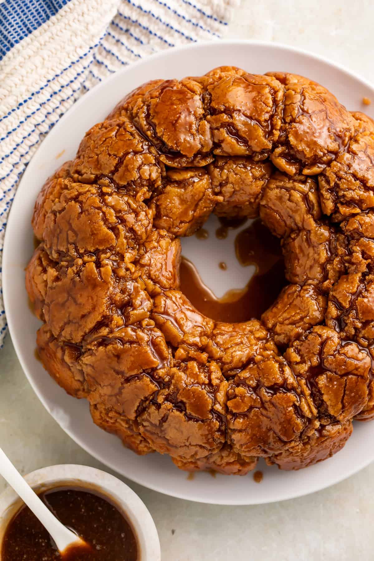 Overhead view of a monkey bread drizzled with caramel on a white plate. A white bowl of caramel with a white spoon sits off to one side.