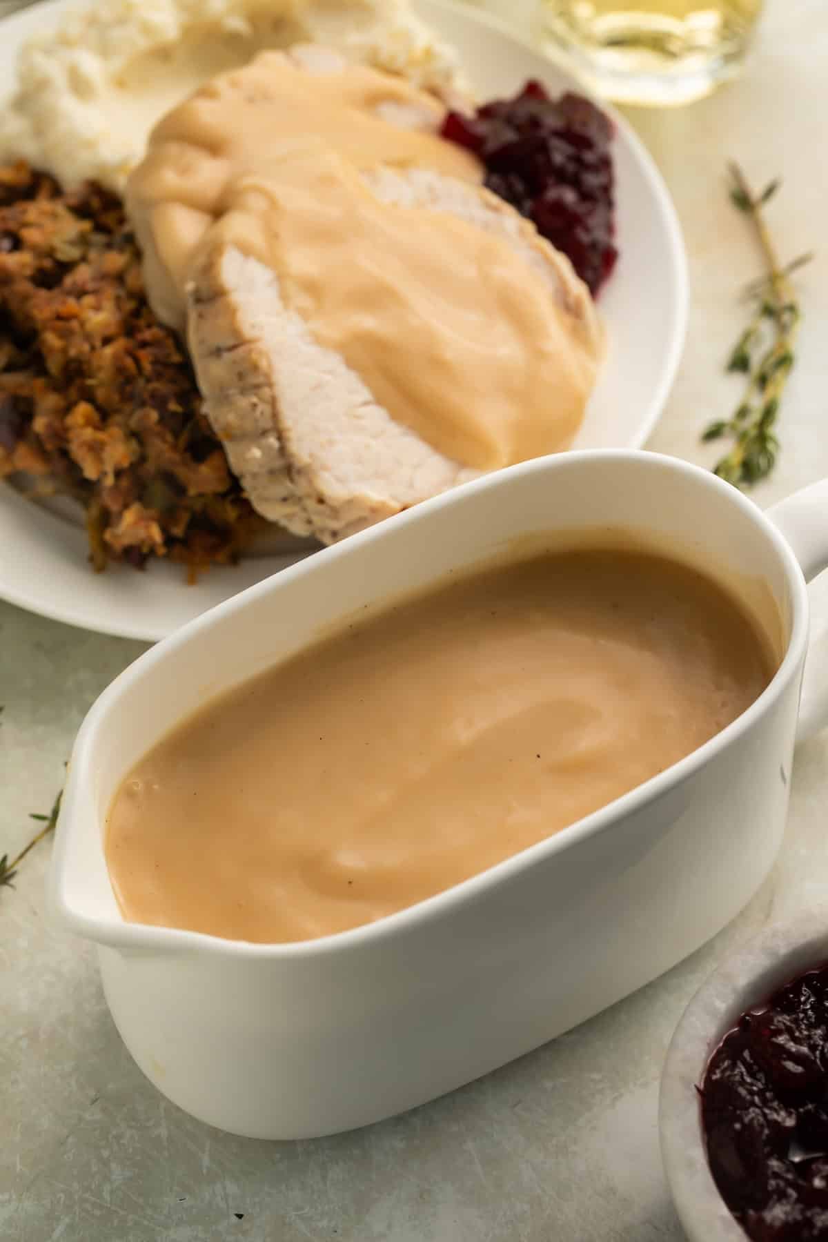 White gravy boat of gravy with gravy-covered turkey and stuffing on a white plate in the background.