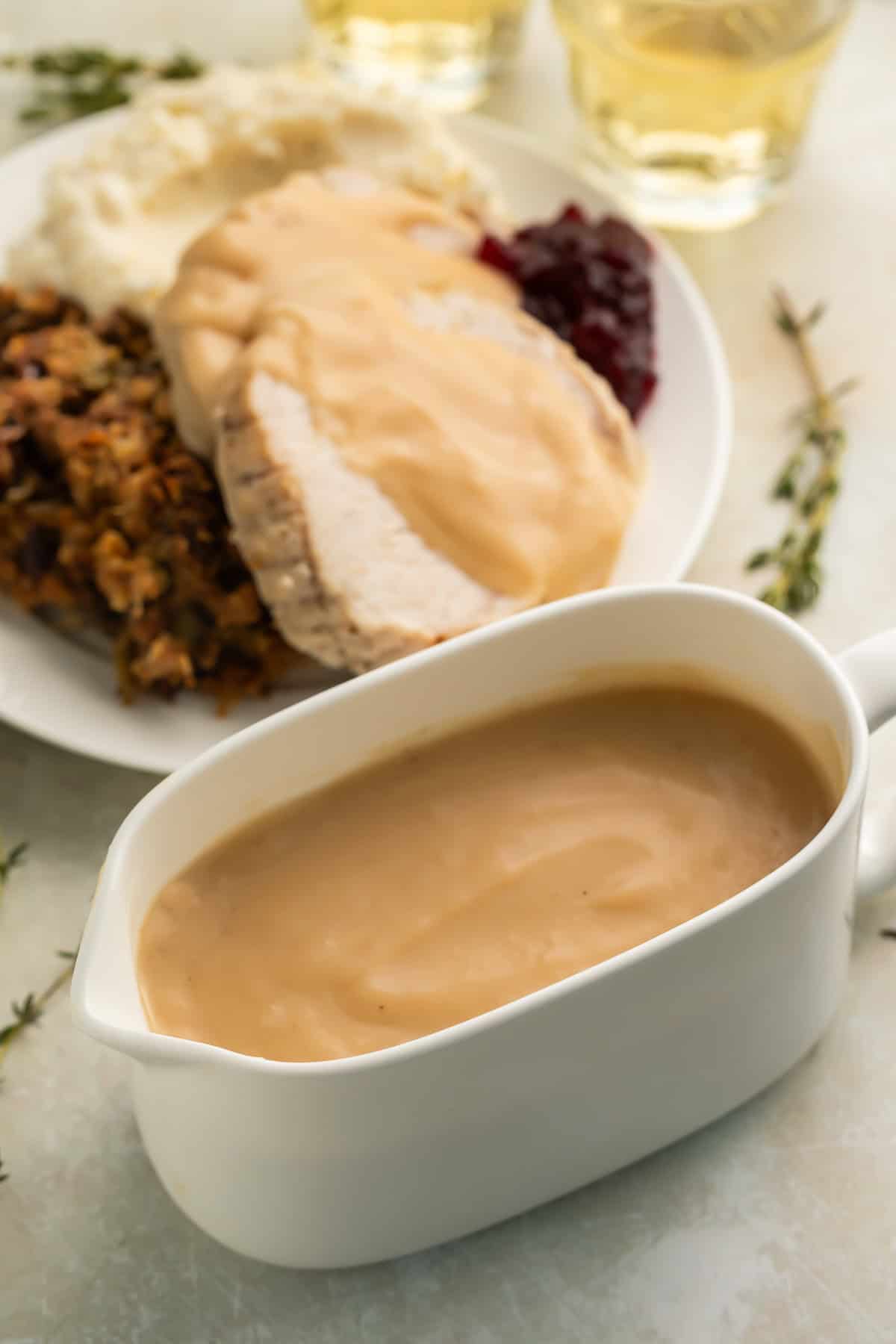 Close up of a white gravy boat with gluten free turkey gravy. There is a thanksgiving dinner with gravy on a plate in the background. 