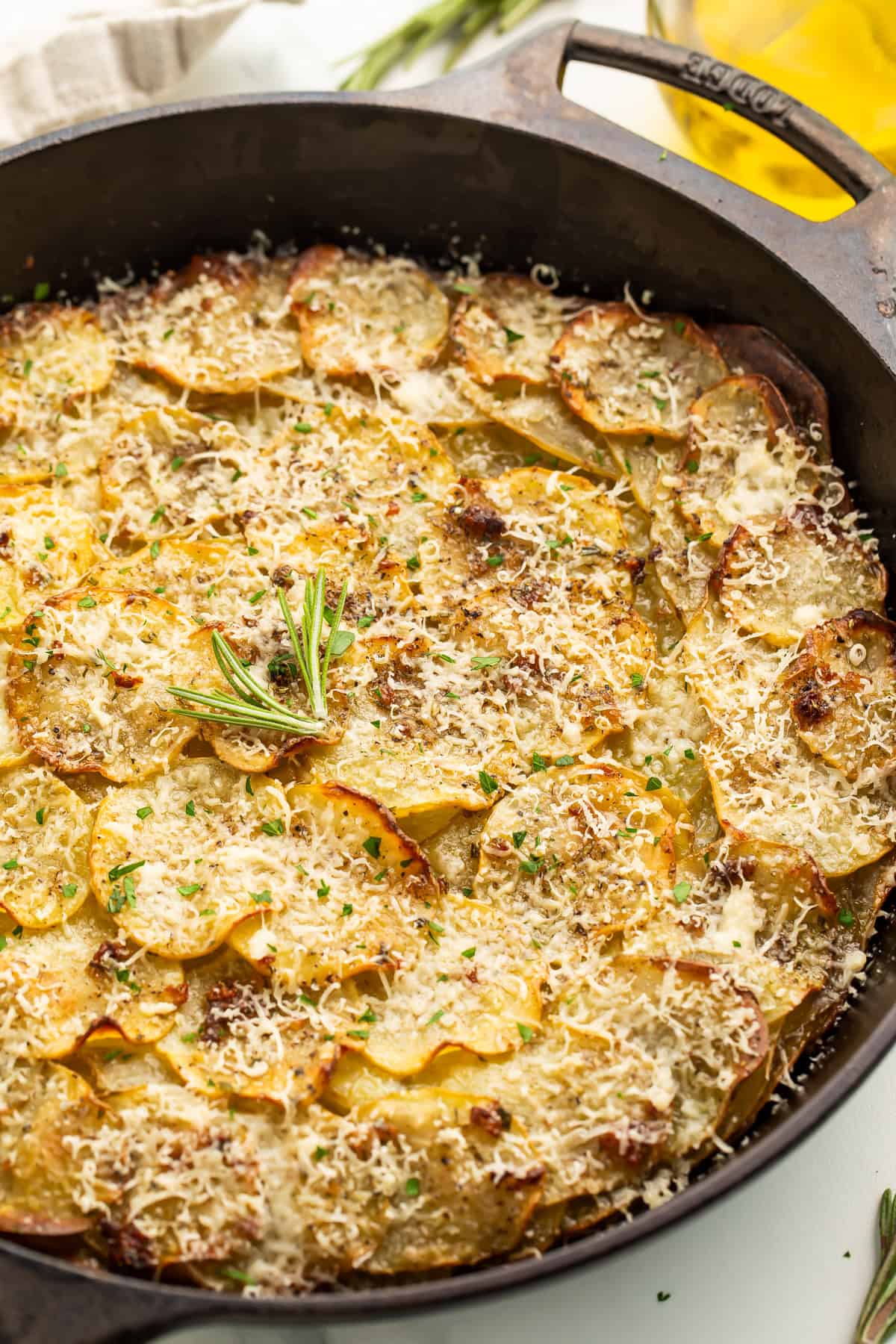 Close up image of a potato galette in a cast iron skillet, covered in melted grated cheese and garnished with a fresh rosemary tip.