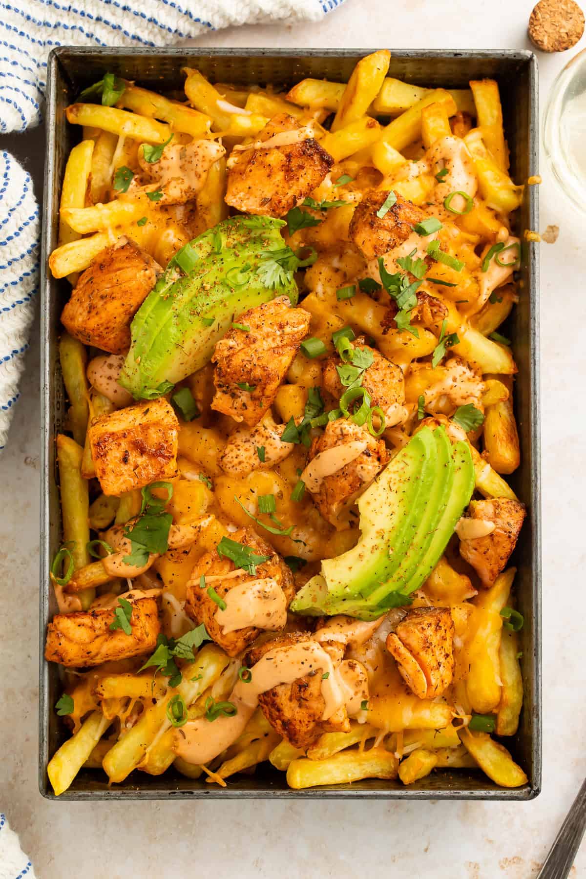Metal tray of cooked fries topped with blackened salmon, creamy sauce and sliced avocado.