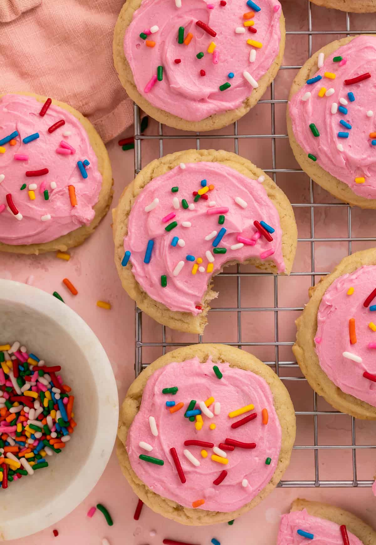 Pink frosted sugar cookies next to a bowl of rainbow sprinkles. One has a bite take out of it.