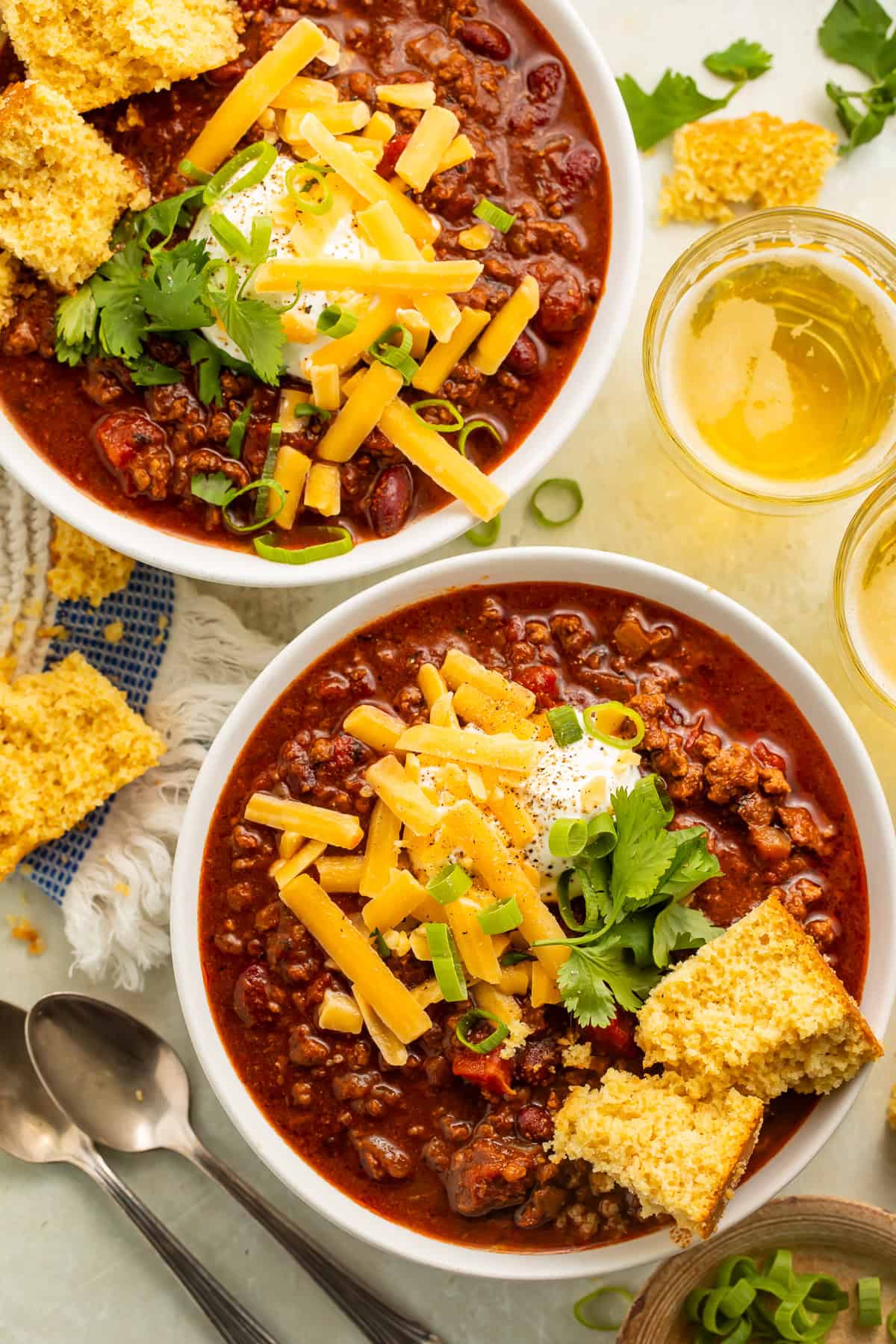 Two white bowls of chili topped with cornbread, green onions, grated cheese and sour cream.