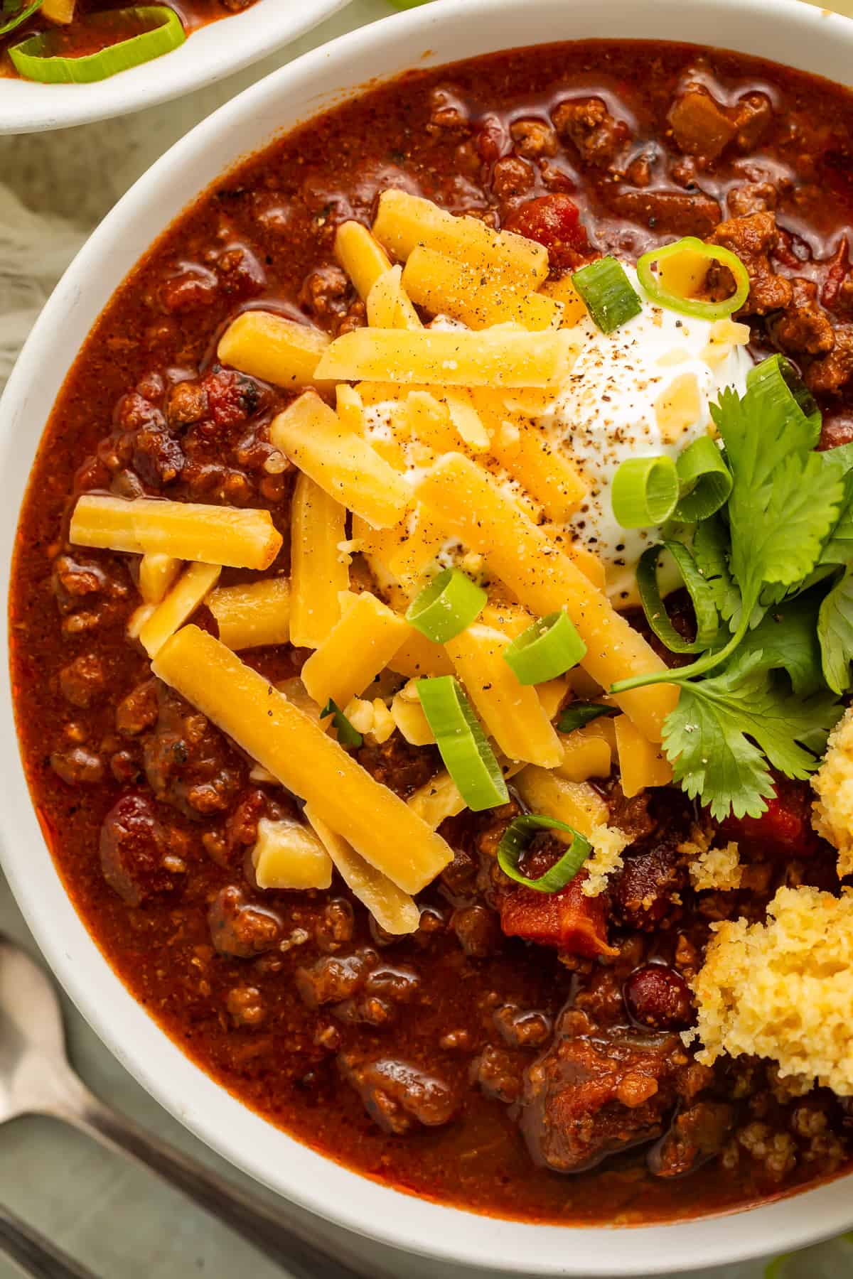 Close up of a bowl of chili with beans and ground meat, topped with shredded cheese, sour cream, green onions, and cilantro.