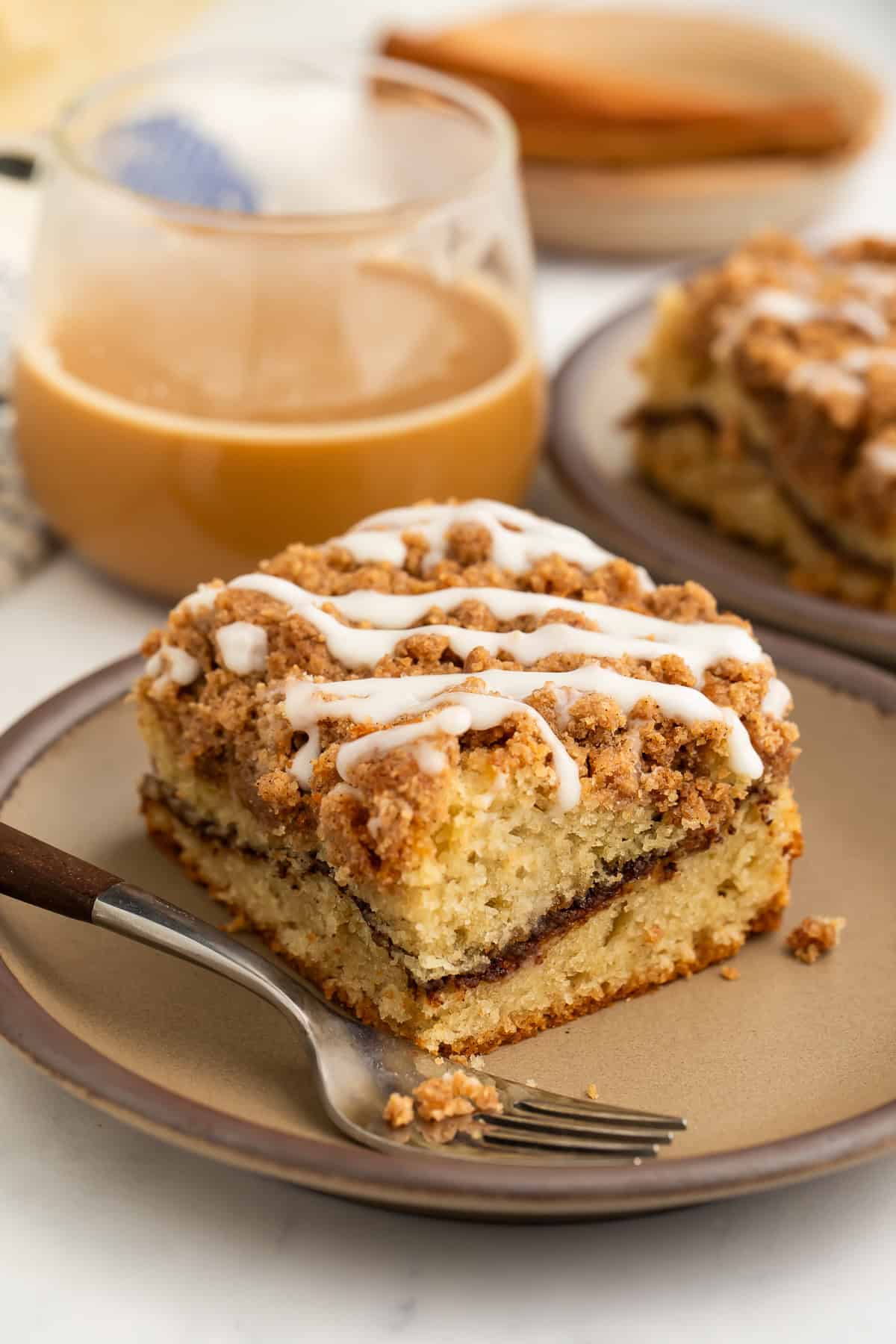 Slice of gluten free coffee cake with streusel topping on a plate with a fork and an iced coffee in the background.