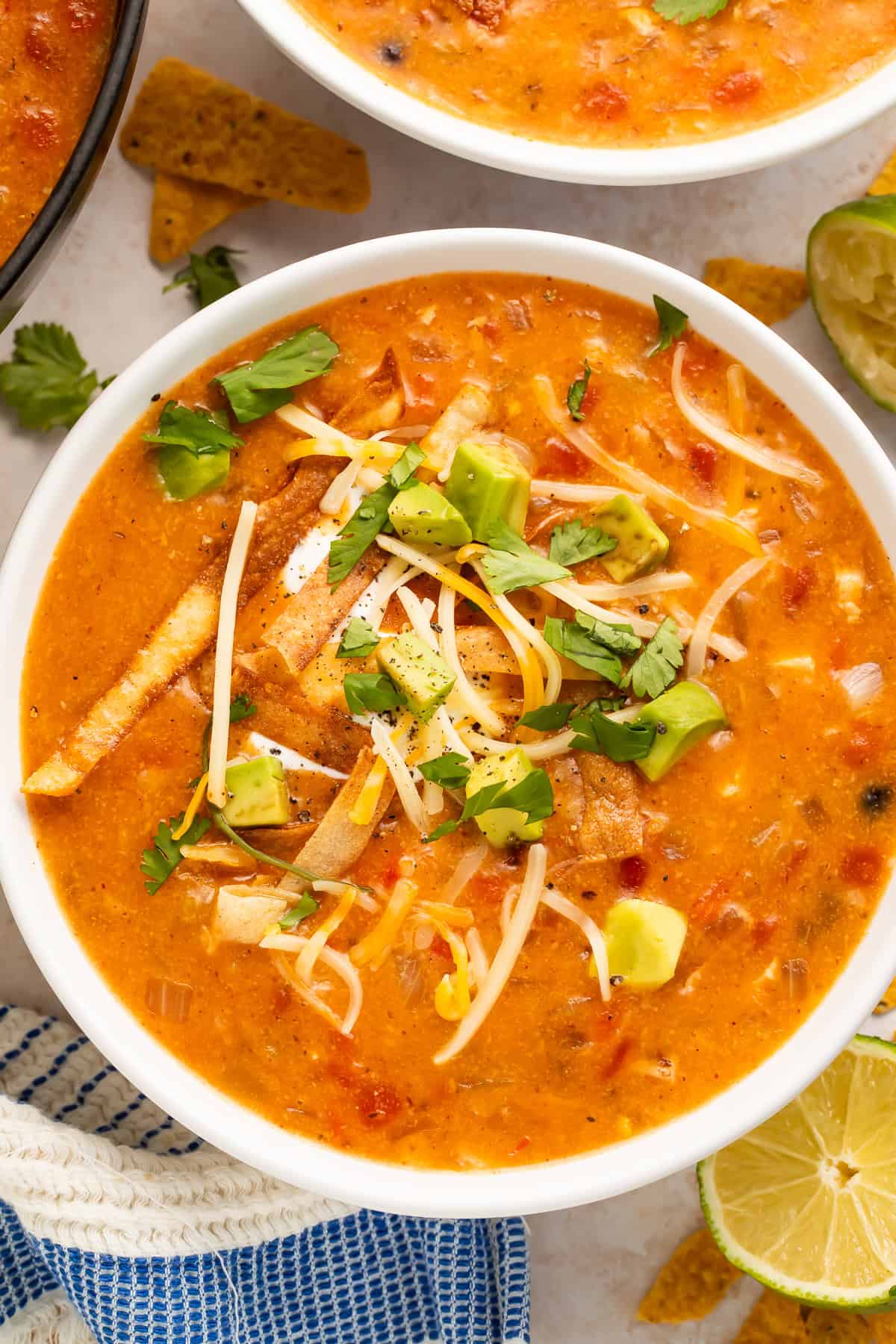 Close up of a white bowl of tortilla soup topped with avocado, shredded cheese and cilantro.
