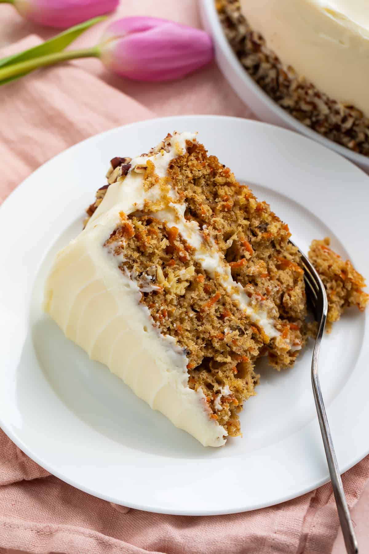 Slice of gluten free carrot cake on a white fork against a pink background with a pair of pink tulips.