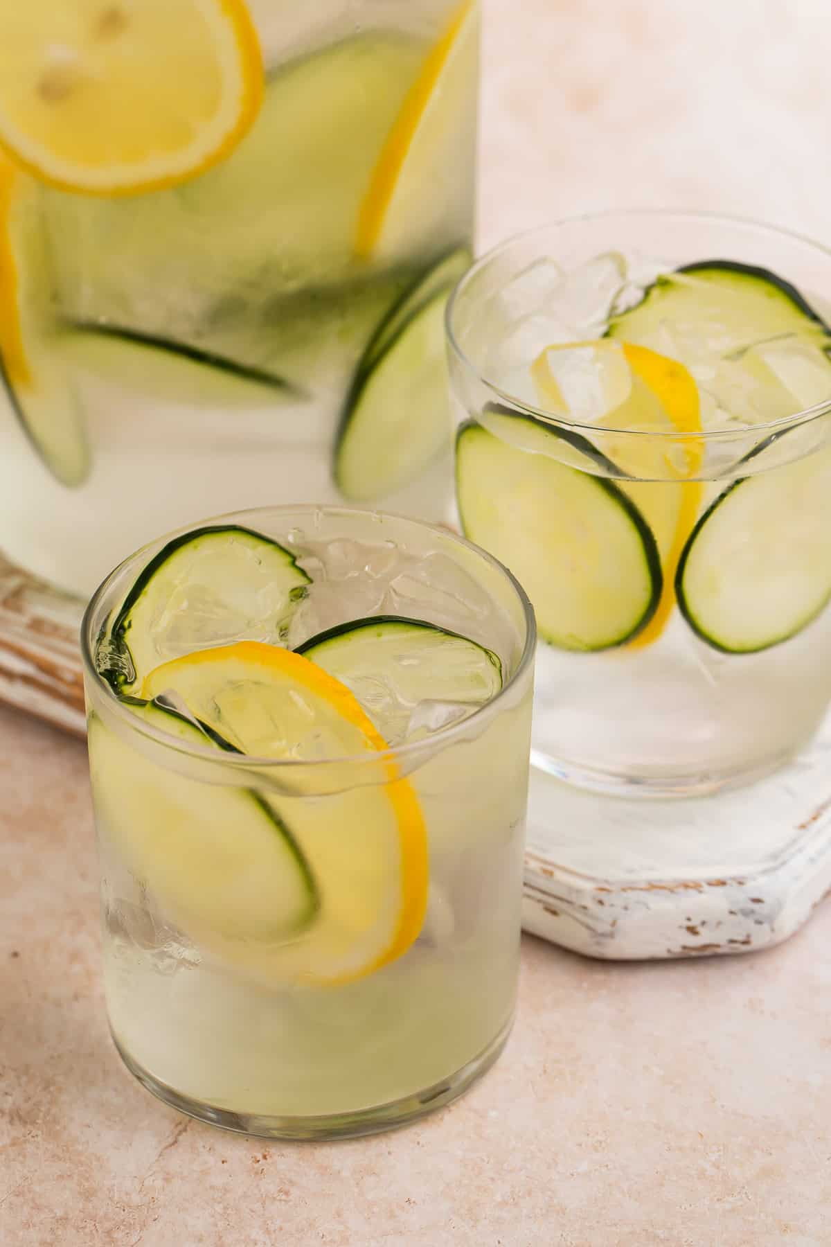 Close up of two glasses of cucumber water with lemon wheels. The jug of infused water is in the background.