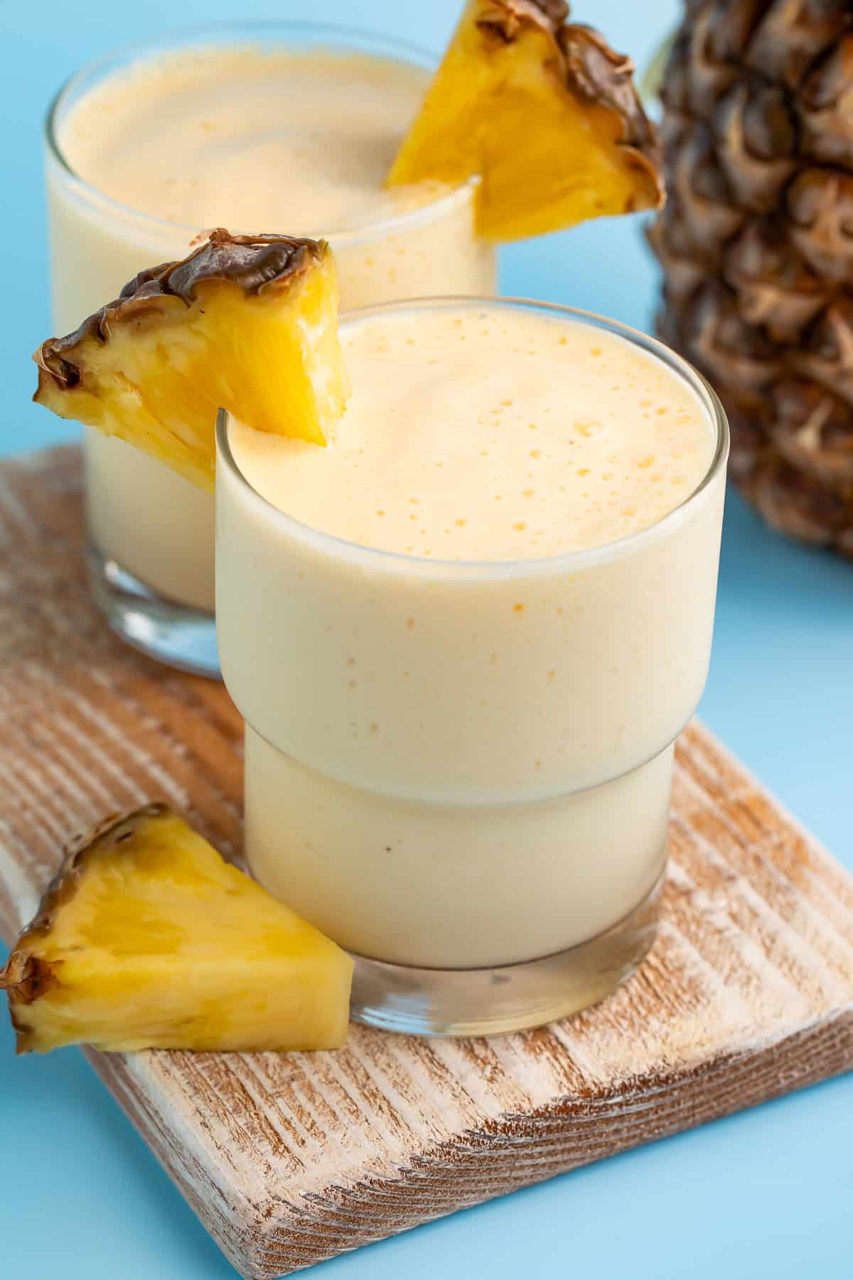 Side-on view of two glasses of tropical smoothie on a wooden board, garnished with fresh pinapple.