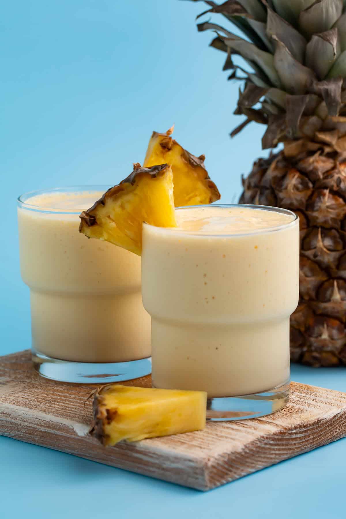 Two glasses of creamy yellow mango and pineapple smoothie on a wooden board against a blue background. A fresh pineapple is behind them and they're garnished with fresh pineapple slices.