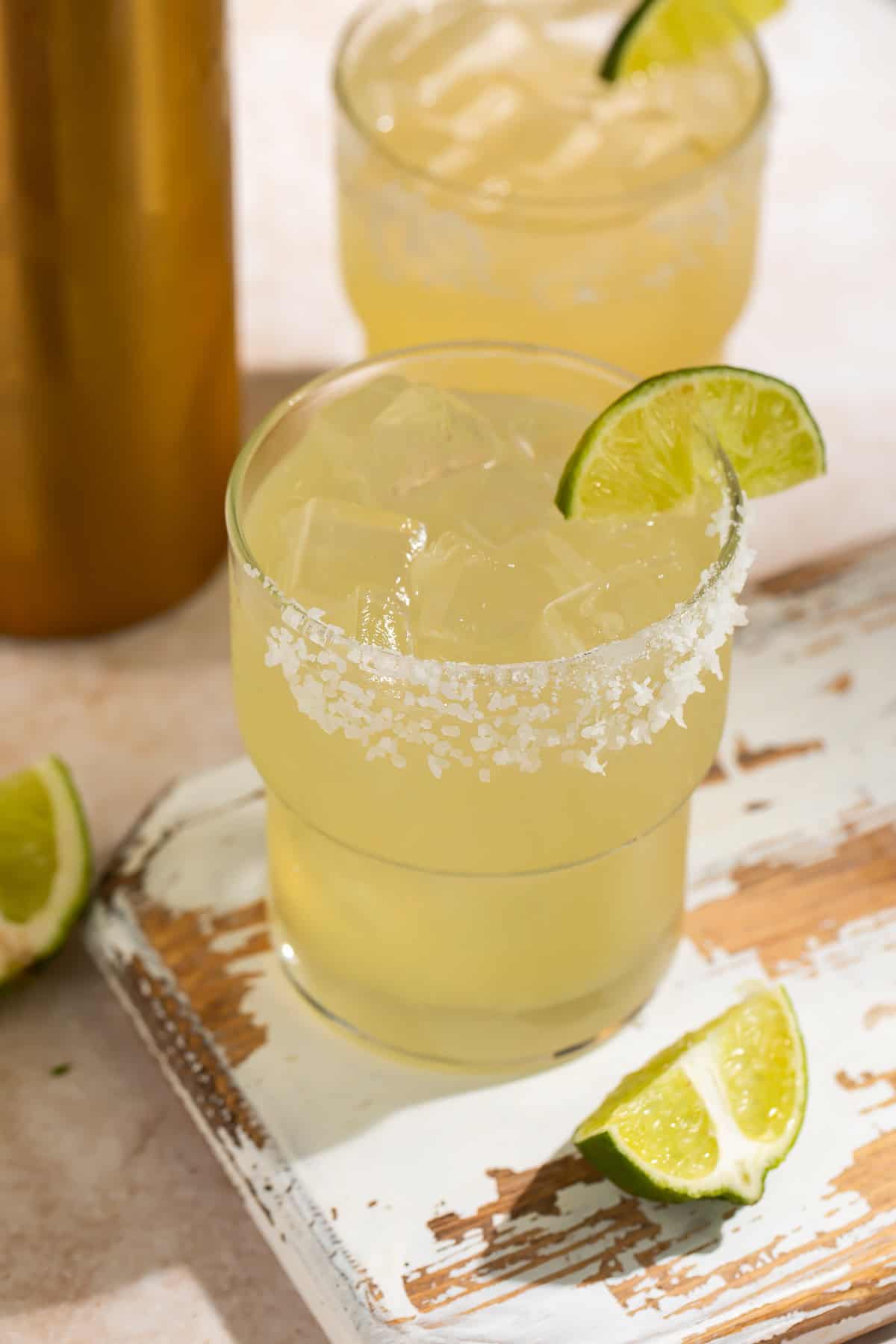 Margarita in a short glass with ice, a salt rim and mile wedges on a wooden board. A cocktail shaker base and another drink are visible in the background.