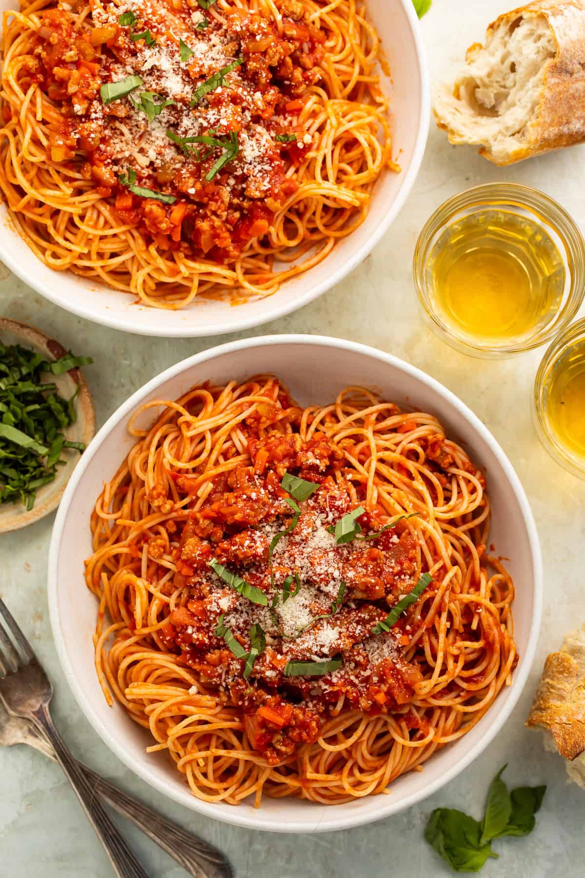 Two bowls of ground turkey spaghetti next to cups of white wine and a torn baguette.