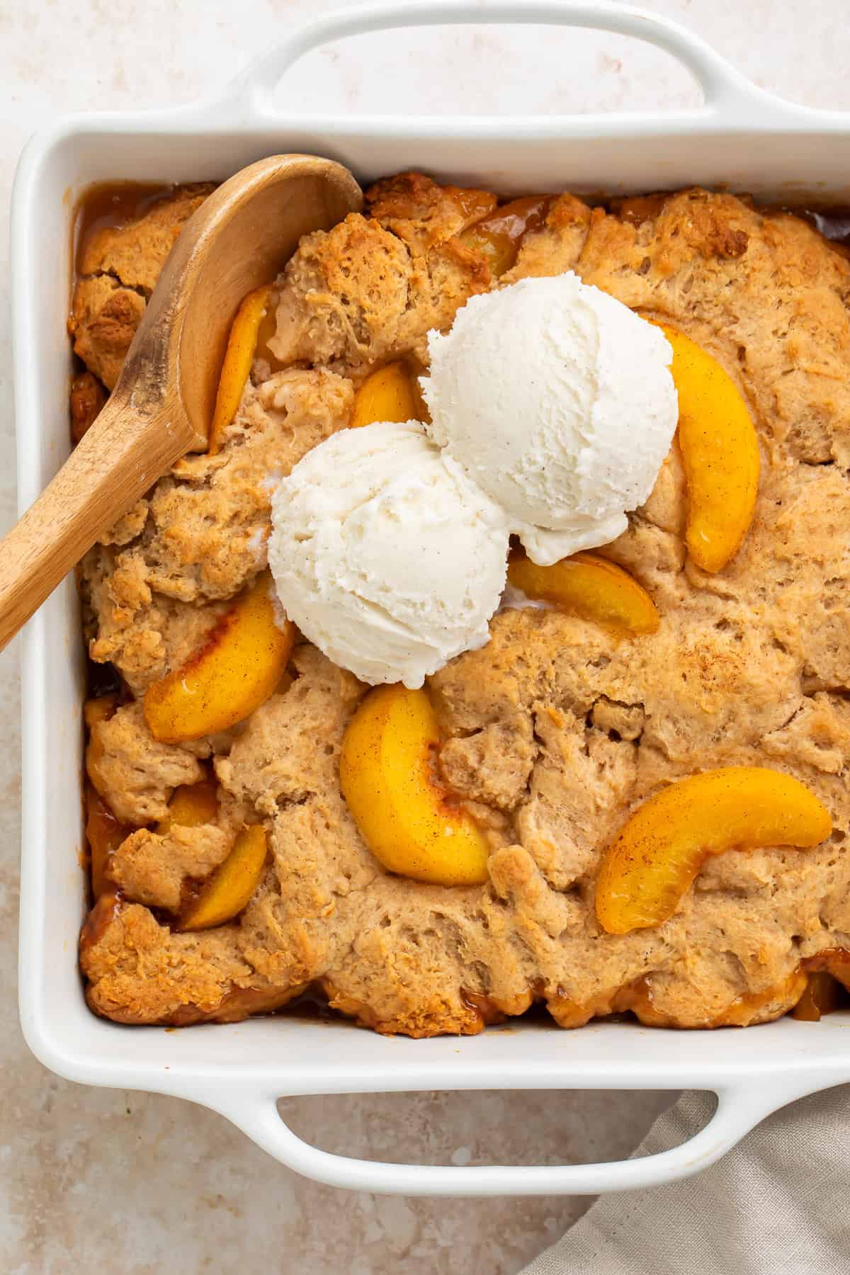 Square baking dish with peach cobbler topped with a scoop of vanilla ice cream.