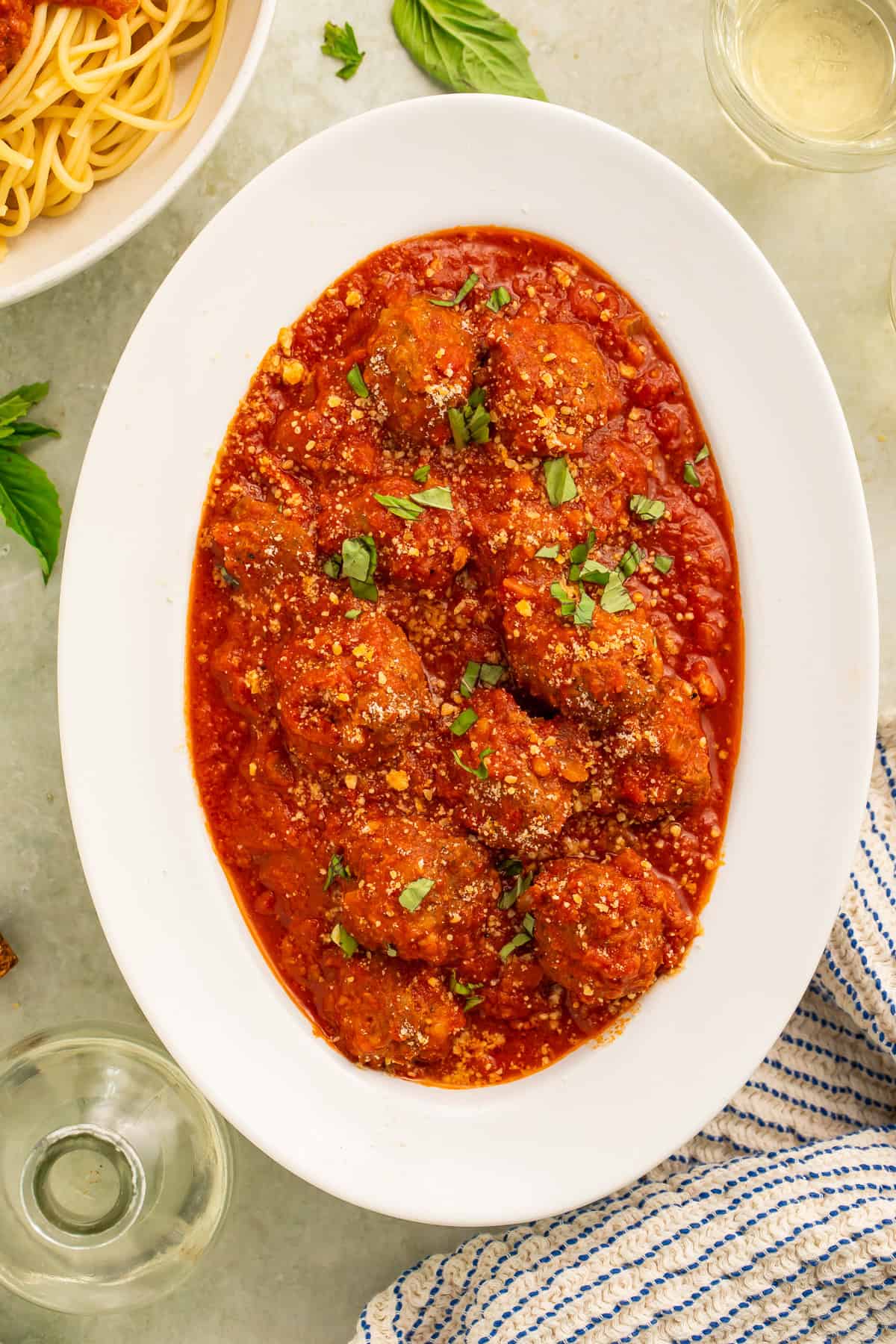 White oval serving platter of meatballs in tomato sauce. A bowl of cooked spaghetti and bottle of olive oil is visible off to one side.