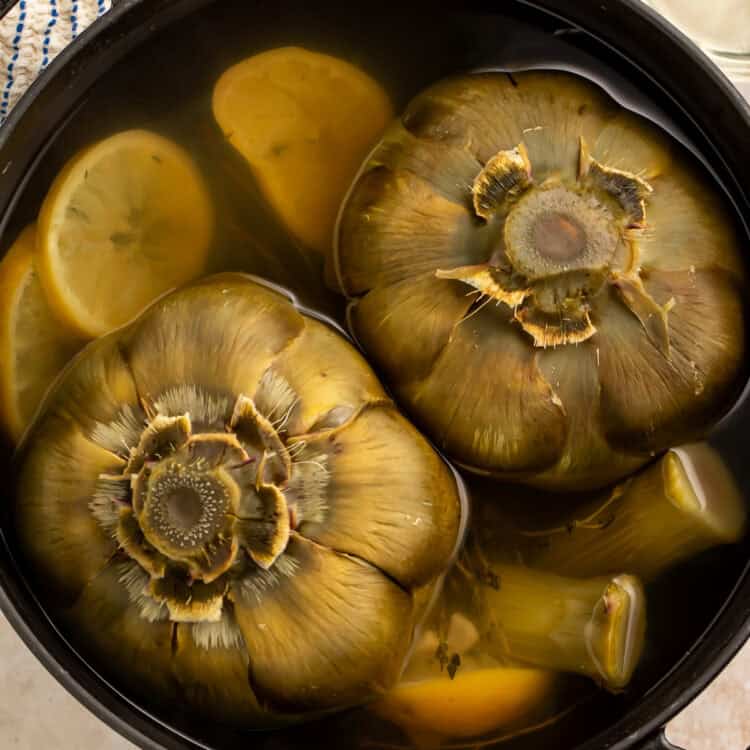 Whole artichokes boiling in a pot with lemon slices and garlic cloves