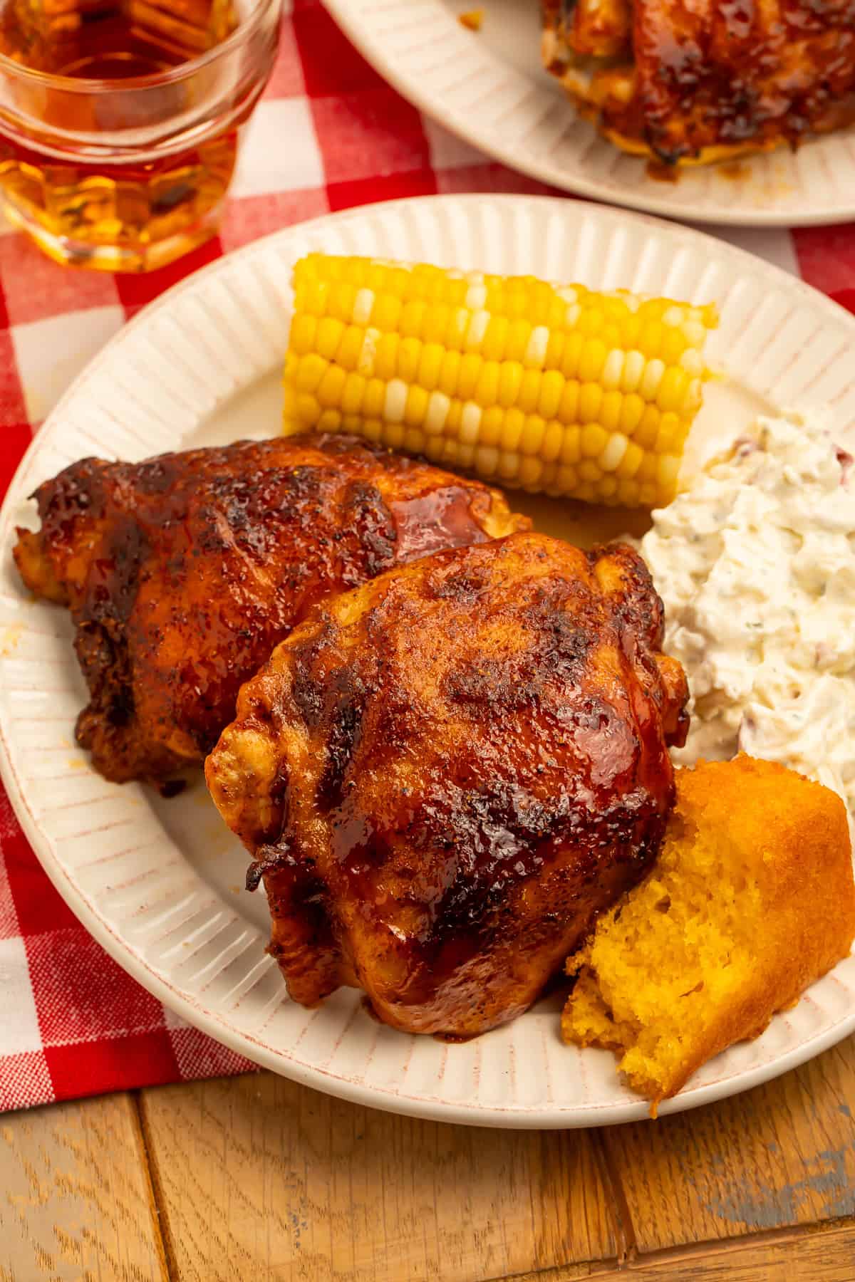 Baked BBQ chicken thighs served with potato salad, corn on the cob, and cornbread on a dinner plate.