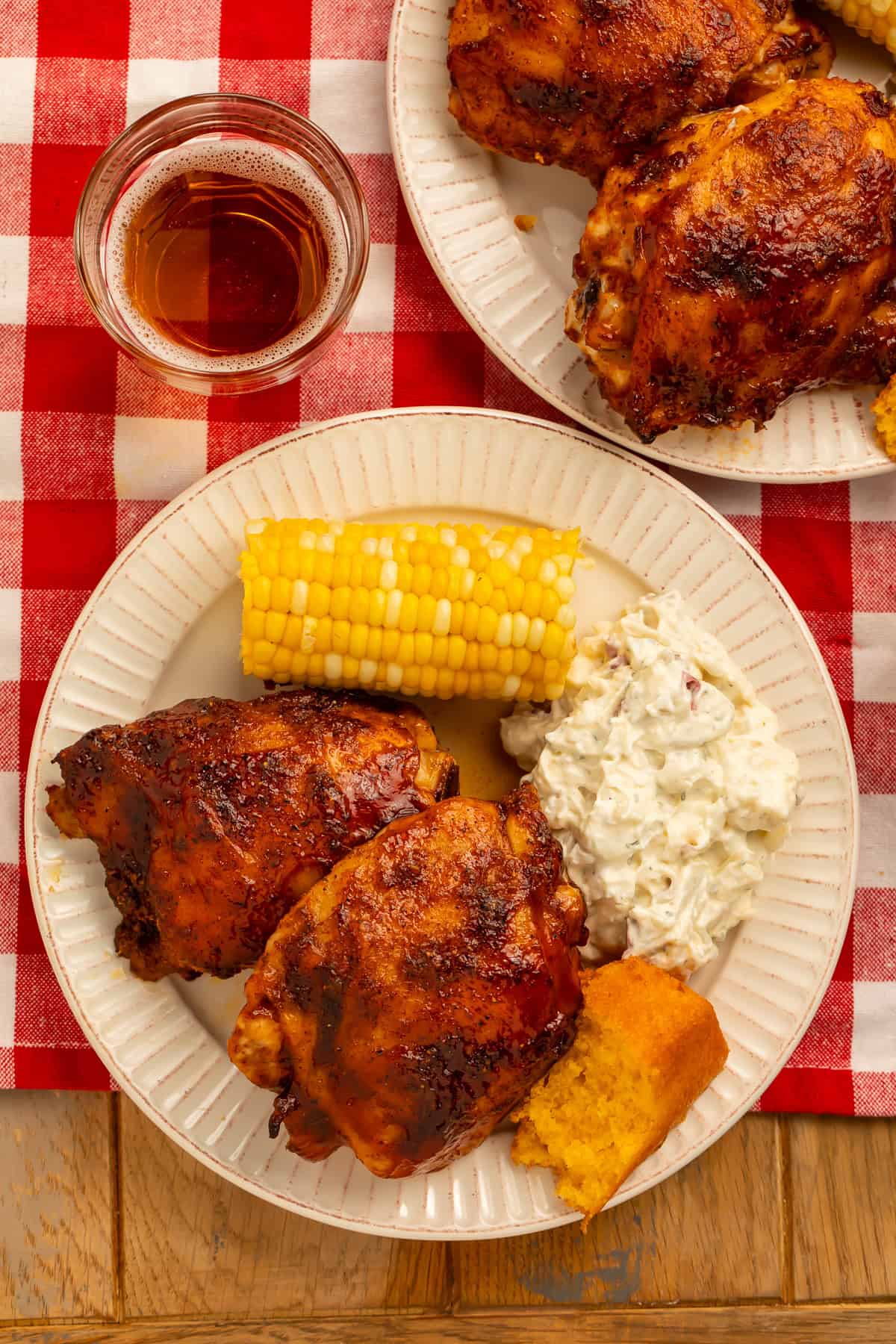 Overhead view of baked BBQ chicken thighs with potato salad, corn on the cob, and cornbread.