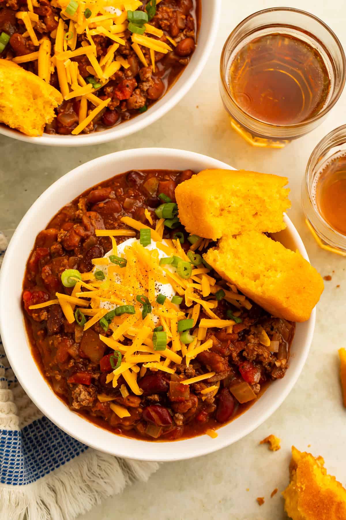 Overhead view of two bowls of bison chili with cheese, sour cream, green onions, and cornbread.