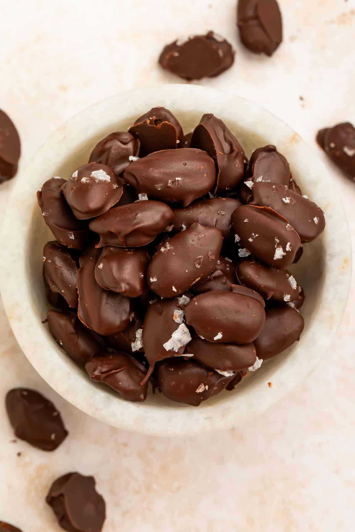 Dark chocolate-covered almonds topped with flaky sea salt in a white bowl.
