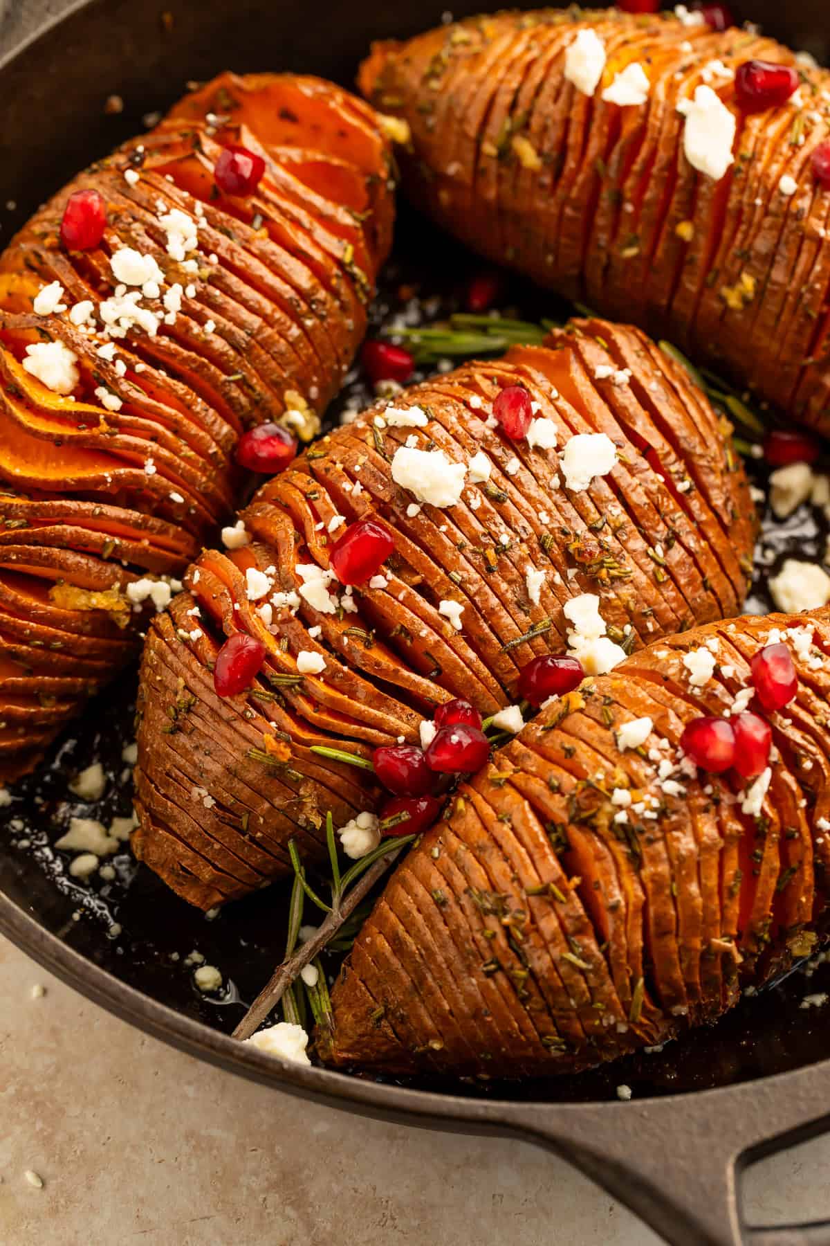 Rosemary maple hasselback sweet potatoes topped with crumbled feta and pomegranate seeds in a cast iron skillet.