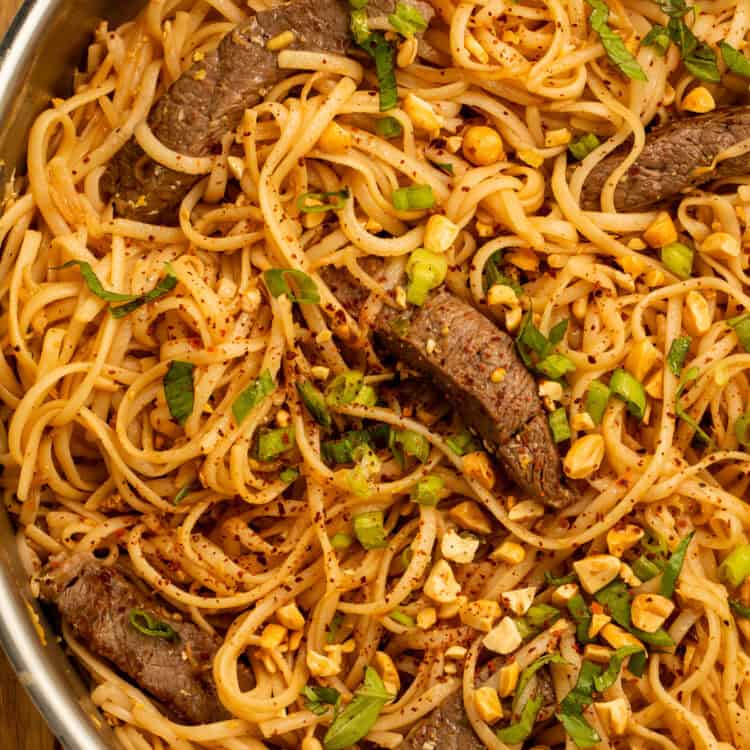 Close-up of beef pad Thai in a skillet with rice noodles, sliced beef, peanuts, green onions, and chili flakes.