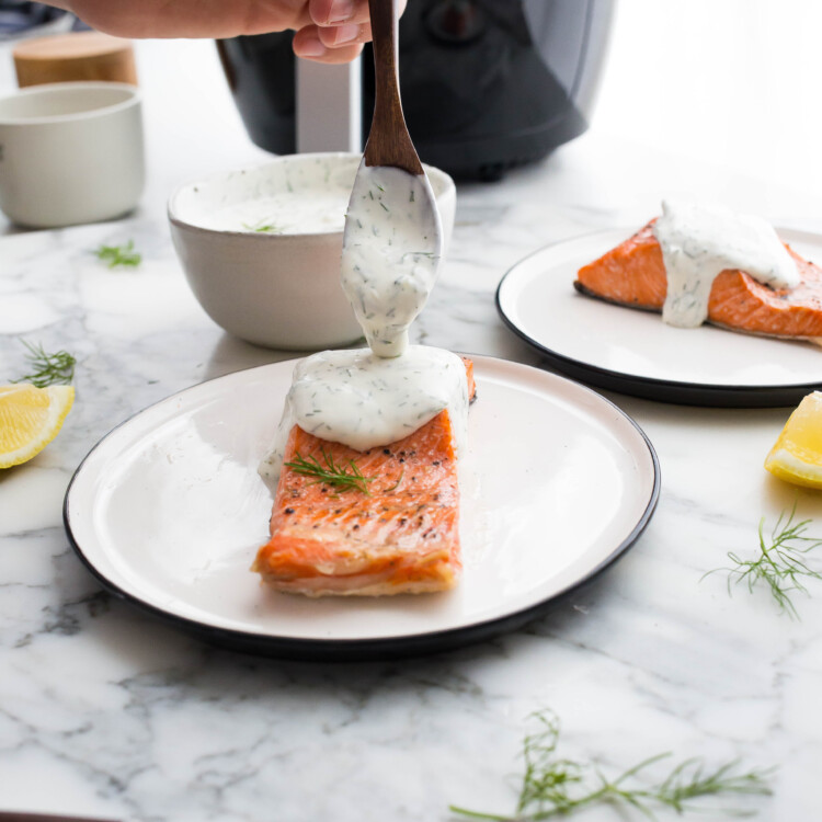 A hand spooning creamy dill sauce on a piece of salmon