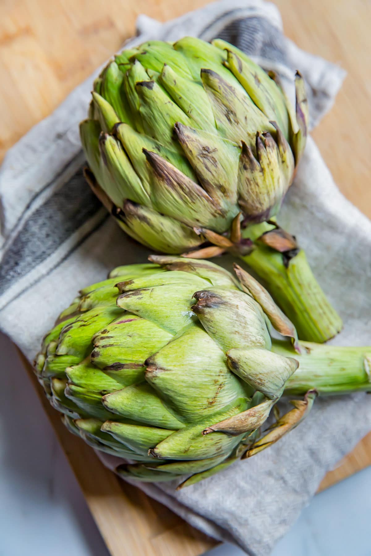 Two fresh, unboiled artichokes on a cutting board with a kitchen towel.