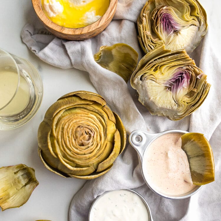 Boiled artichokes on a table with three different dipping sauces.