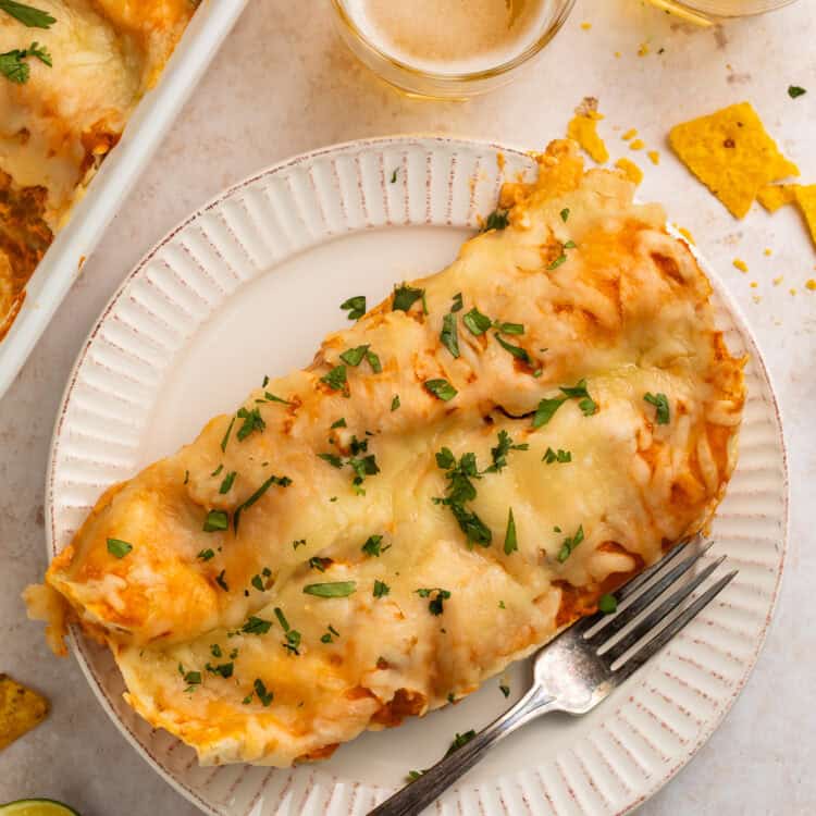 Two chicken enchiladas on a white plate with a form,. There are glasses of beer and the pan of other enchiladas off to one side.