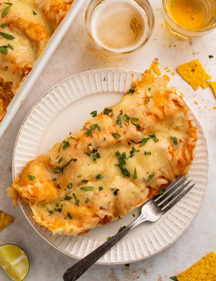 Two chicken enchiladas on a white plate with a form,. There are glasses of beer and the pan of other enchiladas off to one side.