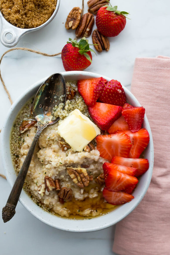 White bowl of oatmeal with strawberries, butter, hemp seeds, pecans, brown sugar, and maple syrup next to pink napkin and spoon.