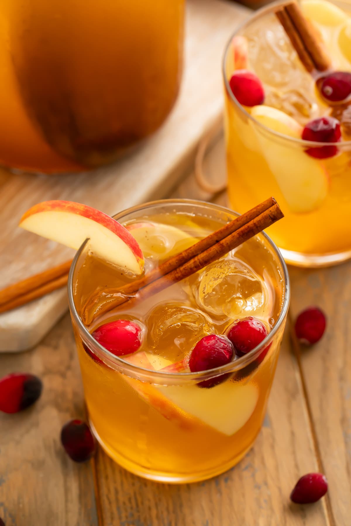Zoomed-out, angled view of a glass of apple cider sangria, in front of a pitcher, garnished with apple slices, grapes, and a cinnamon stick.