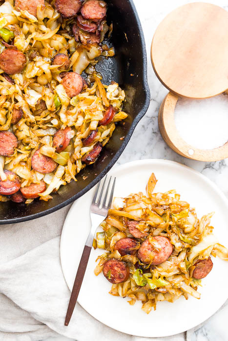 Kielbasa and cabbage in cast iron skillet and white plate with fork and salt.