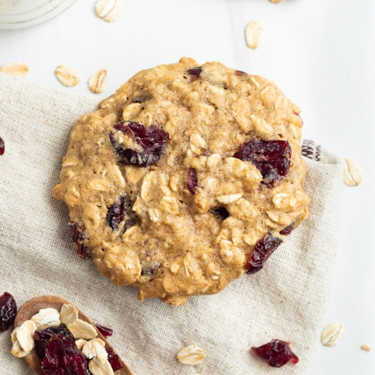 Overhead view of cranberry oatmeal cookie on a napkin next to a wooden spoon with cranberries and oats.