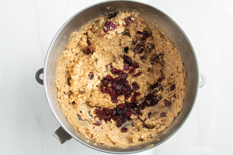 Overhead view of rehydrated cranberries added to a large silver stand mixer bowl containing oatmeal cookie dough.